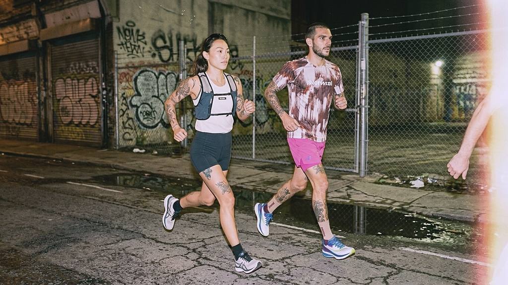 Two runners mid-stride on a city street at night, passing a graffiti wall.